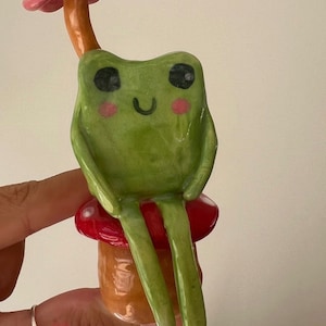 以下が含まれることがあります： ピンクの花の傘を持った緑色のセラミックの蛙が、赤と茶色のキノコの上に座っています。蛙は黒い目、黒い笑顔、そしてピンクの頬をしています。