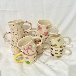 May include: A selection of handmade ceramic mugs with diverse designs. Some mugs display floral patterns, while others feature a pink bow, a frog, or colourful polka dots. The mugs have a cream base and are stacked on a white surface.