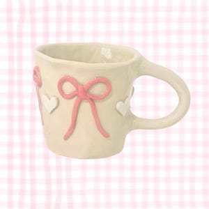 May include: Cream ceramic mug with a curved handle, decorated with pink bows and white hearts. The mug has an irregular shape, giving it a handmade appearance. The background is a pink and white gingham pattern.