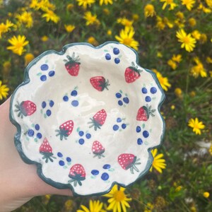 Puede incluir: Un pequeño cuenco de cerámica blanca con borde festoneado, pintado a mano con fresas rojas y arándanos azules. El borde es de color verde azulado oscuro. El cuenco se sostiene frente a un fondo de flores amarillas.