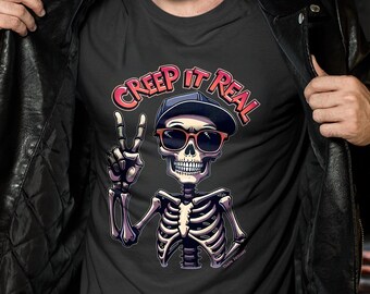 May include: Dark gray t-shirt featuring a skeleton wearing sunglasses and a cap, giving a peace sign. The text "CREEP IT REAL" is arched above the skeleton. The shirt is being held open by a person wearing a black leather jacket.