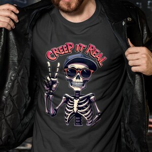 May include: Dark gray t-shirt featuring a skeleton wearing sunglasses and a cap, giving a peace sign. The text "CREEP IT REAL" is arched above the skeleton. The shirt is being held open by a person wearing a black leather jacket.