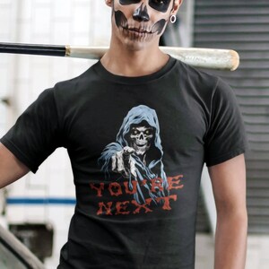 May include: A black t-shirt with a graphic of a hooded figure with a skull face pointing at the viewer. The text "YOU'RE NEXT" is printed below the figure.