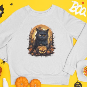 May include: White sweatshirt featuring a Halloween-themed graphic. The design includes a black cat with glowing yellow eyes, a carved pumpkin, and a full moon in the background. The cat is surrounded by pumpkins and autumn leaves.