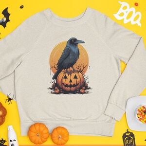 May include: Beige sweatshirt featuring a Halloween-themed graphic. The design includes a black crow perched on a carved orange pumpkin with a smiling face, set against a yellow sun. The sweatshirt is a raglan style.