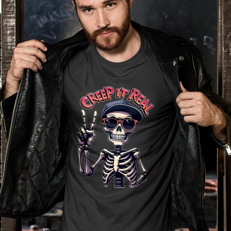 May include: Dark gray t-shirt featuring a skeleton wearing sunglasses and a cap, giving a peace sign. The text "CREEP IT REAL" is arched above the skeleton. The shirt is being held open by a person wearing a black leather jacket.