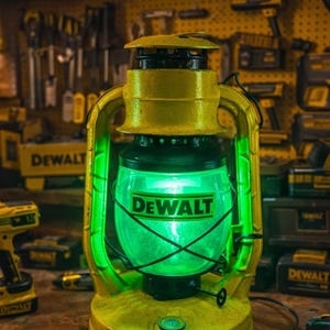 以下が含まれることがあります： 緑色に光る内部を備えた、黄色と黒のDeWaltランタン。ランタンは、ハンドルとベースを備えたクラシックなデザインです。「DEWALT」の文字が前面に印刷されています。背景にはツールとツールボックスが表示されています。