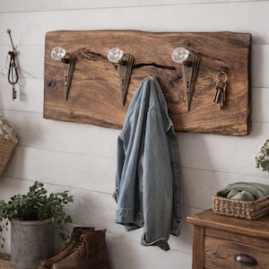 May include: A rustic wooden coat rack with three decorative glass knobs and metal hooks. A denim jacket hangs from one of the hooks. The rack is mounted on a white-painted wood-paneled wall. Other items include a woven tote bag, keys, and a potted plant.