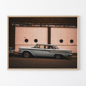 May include: Framed print of a vintage light blue car parked on a street. The car is in front of a building with pink doors and circular details. The image has a vintage aesthetic, evoking a sense of nostalgia.