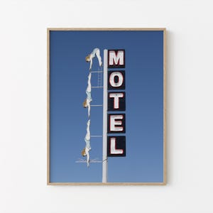 May include: A framed print featuring a blue sky background with a vintage motel sign. The sign spells out "MOTEL" in white letters with red outlines. Three figures in light blue swimsuits are diving from a ladder.