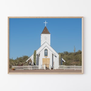 May include: A framed photograph of a white church with a cross on top, set against a clear blue sky. The church has a steeple and a wooden door. Cacti and desert vegetation surround the building.