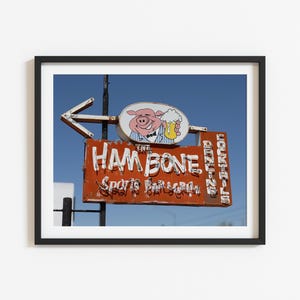 May include: A framed photograph of a weathered, rectangular orange sign for The Ham Bone Sports Bar & Grill. The sign features a cartoon pig holding a beer, a directional arrow, and vertical text reading Dining and Cocktails against a blue sky.