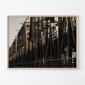May include: A framed photograph of a bridge with a complex metal structure. The bridge is painted in shades of brown and black, with a detailed network of beams and supports. The sky is a pale gray.