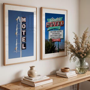 May include: Two framed prints of vintage motel signs. One features the word "MOTEL" with a diving figure, the other reads "Starlite MOTEL" with a list of amenities. Both are in wooden frames, displayed above a wooden table.