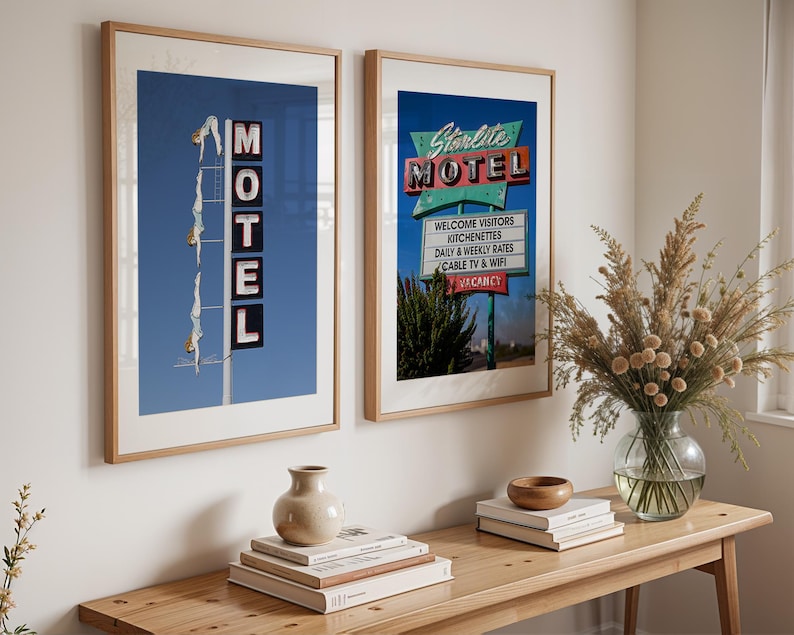 May include: Two framed prints of vintage motel signs. One features the word "MOTEL" with a diving figure, the other reads "Starlite MOTEL" with a list of amenities. Both are in wooden frames, displayed above a wooden table.