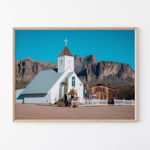 May include: Framed photograph of a white church with a cross on top, set against a backdrop of mountains and a clear blue sky. A wooden building and white fence are also visible. The image is framed in a light brown wooden frame.