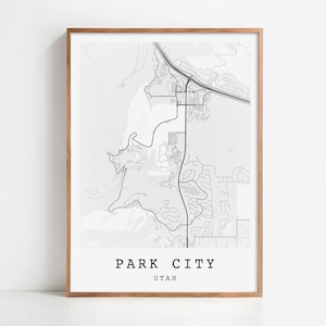 May include: A framed black and white map print of Park City, Utah. The map features roads, buildings, and natural features. The text "PARK CITY UTAH" is printed in a bold, sans-serif font at the bottom of the print. The frame is a light brown color.