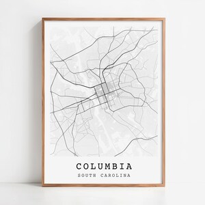 May include: A framed black and white map print of Columbia, South Carolina. The map features a detailed street layout with the city name and state printed in a simple, modern font at the bottom. The print is encased in a light brown wooden frame.