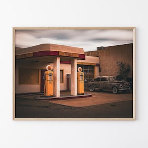 May include: A framed print of an old Shell gas station with two yellow gas pumps. The building is beige with a red and white trim. A vintage car is parked nearby under a cloudy sky.
