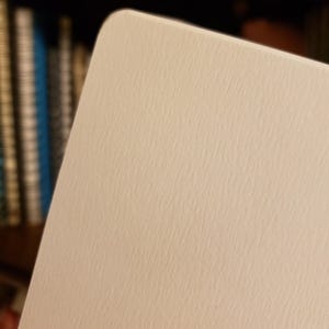 May include: Close-up of a rectangular, off-white card with rounded corners. The card has a textured surface, and the focus is on the upper right corner. The background is blurred, with hints of books on a shelf.