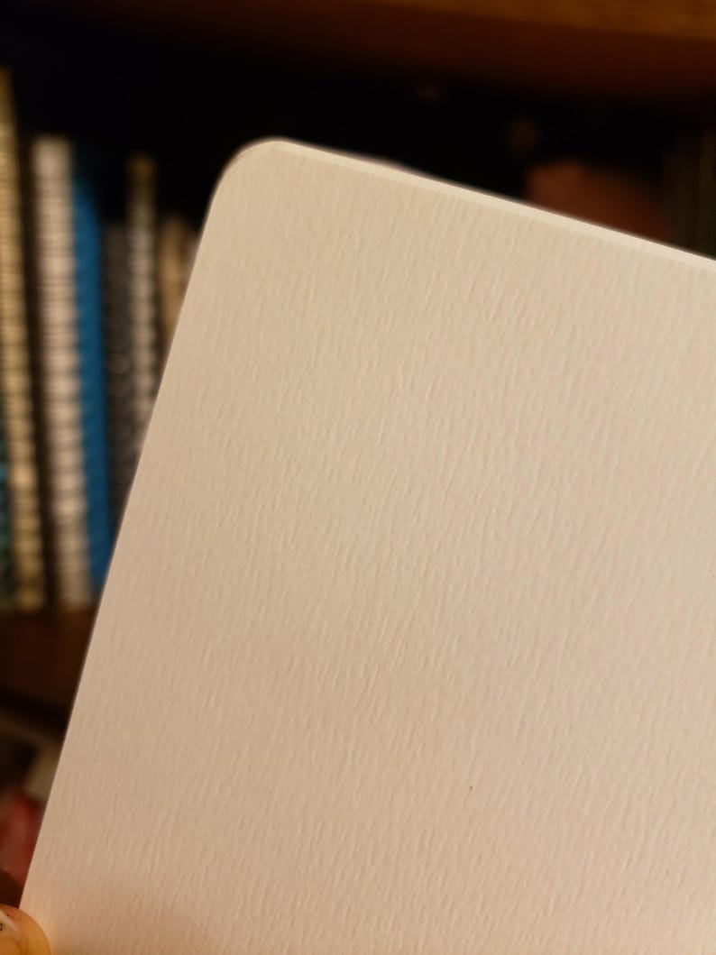 May include: Close-up of a rectangular, off-white card with rounded corners. The card has a textured surface, and the focus is on the upper right corner. The background is blurred, with hints of books on a shelf.
