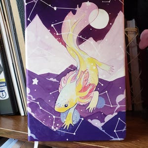 May include: A notebook with a colorful illustration of an axolotl against a purple night sky background. The axolotl is yellow and white with pink gills. The background features mountains, stars, and constellations.