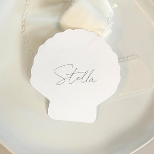 May include: A white shell-shaped place card with the name Stella printed in black script, resting on a white plate with a piece of cream-coloured frayed silk ribbon draped behind the card, creating a clean and simple table setting element.