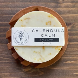 May include: A round, white face soap bar with yellow flecks, labeled "Calendula Calm Face Soap 3+ oz" in black text on a white label. The soap is sitting on a brown, circular soap dish.