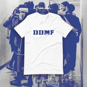 May include: White t-shirt with the blue text "DDMF" in a block font. The shirt is in front of a group of basketball players in a hallway. The players are wearing blue and white uniforms.