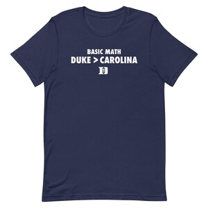 Puede incluir: Camiseta azul marino con texto blanco "BASIC MATH DUKE > CAROLINA" y un logotipo de la Universidad de Duke. La camiseta es de cuello redondo y manga corta.