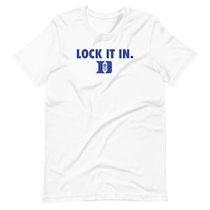 May include: White t-shirt featuring the blue text "LOCK IT IN." and a blue and white emblem. The shirt is made of a soft material and has short sleeves.