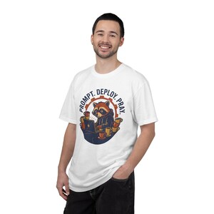 Puede incluir: Una camiseta blanca con un gráfico de un mapache con sudadera trabajando en un portátil, rodeado de tazas de café. El texto PROMPT. DEPLOY. PRAY. está impreso en un arco circular sobre el mapache, con un fondo en forma de engranaje.