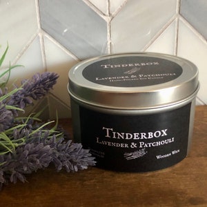 May include: A silver tin candle with a black label that reads "Tinderbox Lavender & Patchouli". The candle has a wooden wick and is next to a sprig of lavender. The background is a white, gray, and blue tile.