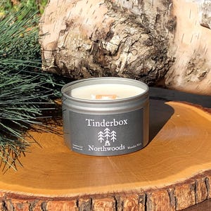 May include: A silver tin candle labeled "Tinderbox Northwoods Wooden Wick" with a white wax interior. The candle sits on a wooden slice, with a birch log and pine needles in the background.