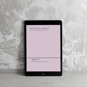 Puede incluir: Una tableta negra que muestra una pantalla rosa con el texto "PORTION & PROMISE DAILY BIBLE READING JOURNAL" y "BOOK 3 PROVERBS THROUGH HABAKKUK". La tableta está sobre un fondo gris texturizado.