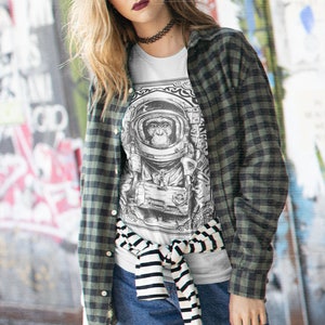 May include: A woman wearing a white t-shirt with a black and white graphic of a monkey in a spacesuit. She is wearing a green and black plaid shirt over the t-shirt and blue jeans. The t-shirt is tied at the waist with a black and white striped belt.