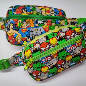 May include: Two fanny packs with green straps and zippers. The larger pack features a comic book print with superheroes like Superman and Batman. The smaller pack has a similar design with cartoon superhero characters. Both packs are on a white background.