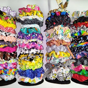 May include: Four vertical stacks of fabric hair scrunchies in a wide variety of colors and patterns, including floral, animal print, polka dot, geometric, and solid designs. The scrunchies are arranged on four black circular bases.
