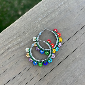 May include: A pair of silver hoop earrings adorned with colorful beaded flowers. The flowers are in shades of red, yellow, blue, green, purple, and white. The earrings are set against a wooden background.