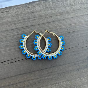 May include: A pair of gold hoop earrings with blue and white beaded flowers. The earrings are on a wooden surface.