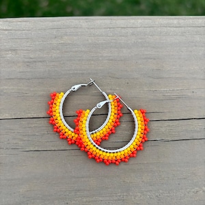 May include: A pair of silver hoop earrings adorned with a vibrant pattern of small, colorful beads. The beads transition from red to orange and yellow, creating a gradient effect. The earrings are displayed on a wooden surface.