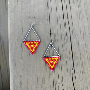 May include: A pair of handmade earrings featuring a geometric design. Each earring has a silver-colored triangular frame with a beaded triangle in red and yellow. The earrings are displayed on a wooden surface.