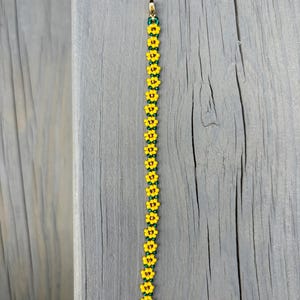 May include: A beaded bracelet featuring a repeating pattern of yellow flower beads with green centers and accents. The bracelet has a gold-colored clasp and is displayed on a gray wooden surface. The floral design gives the bracelet a summery feel.