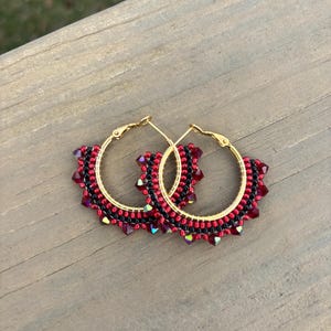 May include: A pair of gold hoop earrings with intricate beadwork. The earrings feature a pattern of red and black beads, with a row of sparkling red crystals along the bottom edge. The earrings are displayed on a wooden surface.