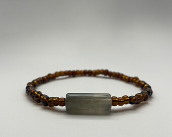 Puede incluir: Una pulsera de cuentas con pequeñas cuentas de color marrón oscuro y una cuenta central rectangular gris. La pulsera está diseñada para ser usada alrededor de la muñeca, sobre un fondo blanco.