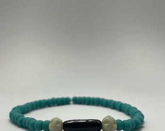 Puede incluir: Un brazalete de cuentas azul turquesa con dos cuentas blancas y una cuenta negra en el centro.