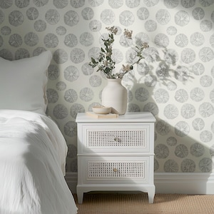May include: A white nightstand with two drawers featuring woven cane fronts. A white vase with dried flowers and a small bowl sit on top. The background is a white wall with a gray citrus slice pattern.