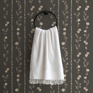 May include: A white, fringed towel hangs from a black circular towel ring. The towel is displayed against a dark gray wall with a floral pattern in a lighter beige color. The towel has a textured design.