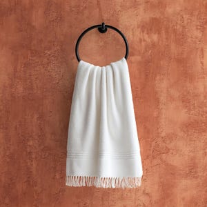 May include: A white, fringed towel hangs from a black metal towel ring. The towel has a textured surface and decorative horizontal lines near the bottom. The background is a textured, reddish-brown wall.