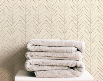 May include: A stack of folded, light gray towels rests on a small white stool. The background features a cream-colored wall with a repeating pattern of gray, chevron-like brushstrokes, creating a textured, neutral aesthetic. The scene evokes a sense of calm and simplicity.